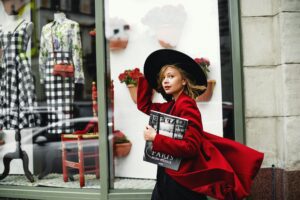 Fashionable woman in a red coat walks by a stylish boutique window, carrying a magazine.