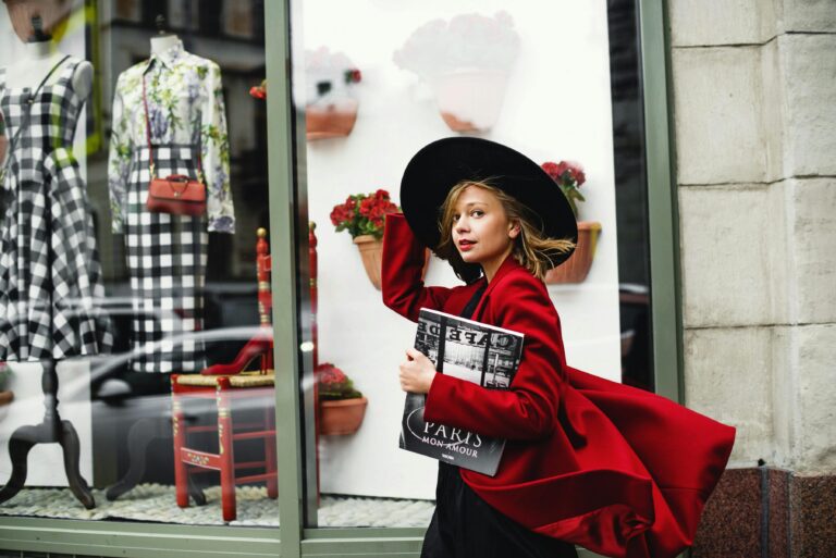 Fashionable woman in a red coat walks by a stylish boutique window, carrying a magazine.