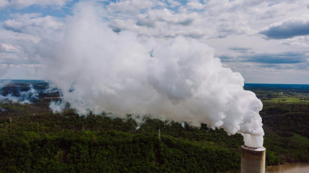 Drone shot of a power plant chimney emitting thick white smoke over a forested landscape.