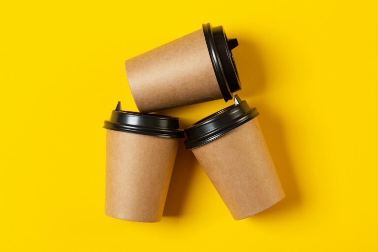 Three eco-friendly paper coffee cups with black lids on a vibrant yellow background representing consumerism lifestyle.
